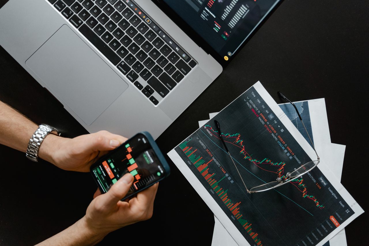 services-02 Hands using smartphone beside laptop with stock charts, showcasing digital trading.