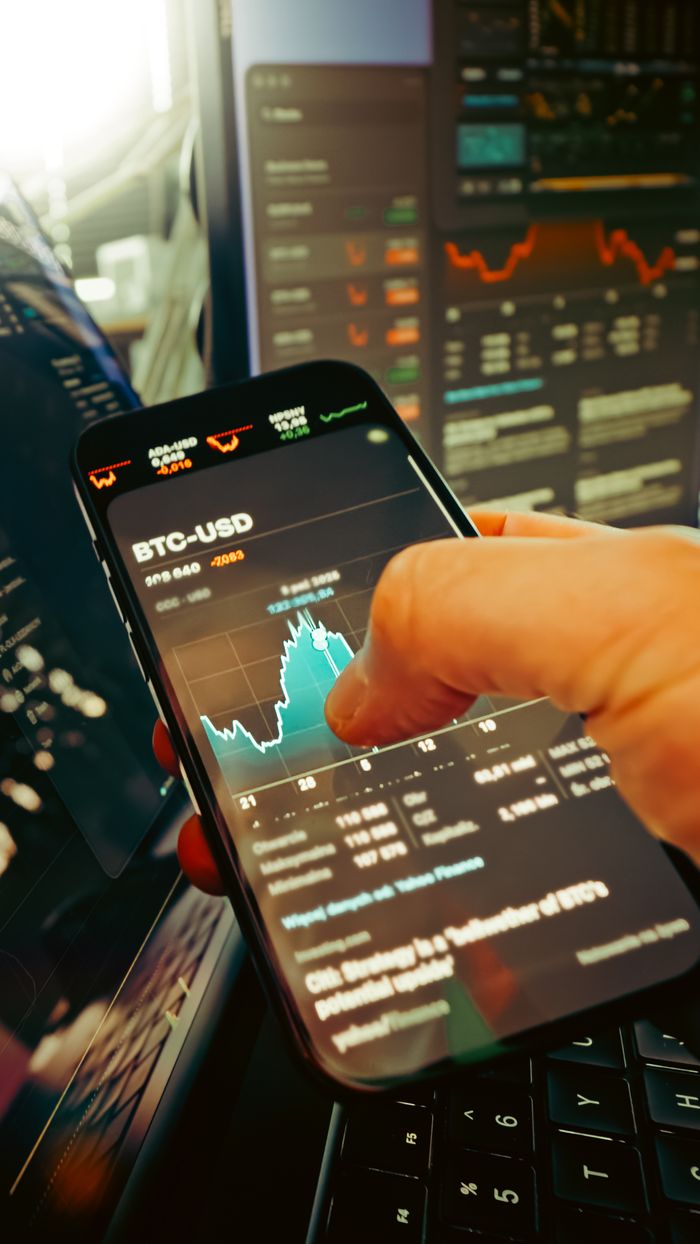 gallery-1 A hand holds a smartphone displaying BTC-USD chart amidst digital trading screens, highlighting crypto finance.